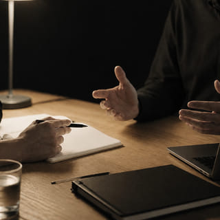 Entretien à deux autour d’une table de travail, éclairé par une lampe.